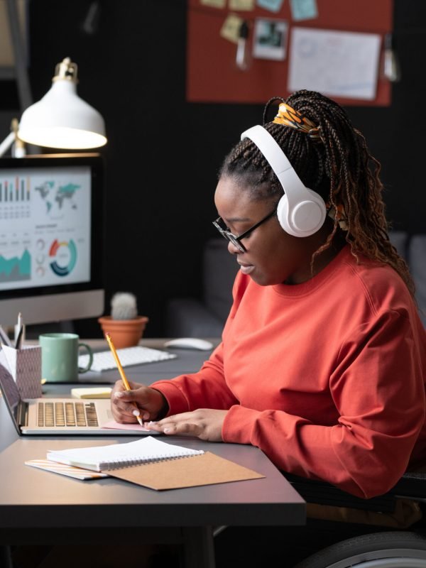 African girl in wireless headphones sitting at the table in front of the laptop and making notes, she studying at home